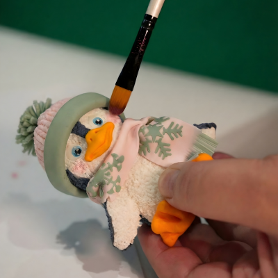 Detailný pohľad na ruku držiacu precízne vymodelovanú cukrovú postavičku tučniaka. Tučniak má realistickú štruktúru povrchu, má oblečenú ružovo-zelenú zimnú čiapku s brmbolcom a ružový šál so zelenými snehovými vločkami. Zhora je priložený kozmetický štetec, ktorým sa práve nanáša ružová púdrová farba na líčko postavičky. Ukážka techník modelovania, textúrovania a tieňovania jedlými farbami z cukrárskych kurzov Artmeli.